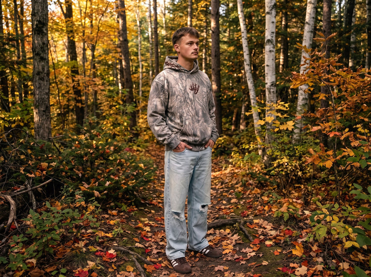 Man in a Covenant Camo hoodie standing on a leaf-covered forest trail in autumn